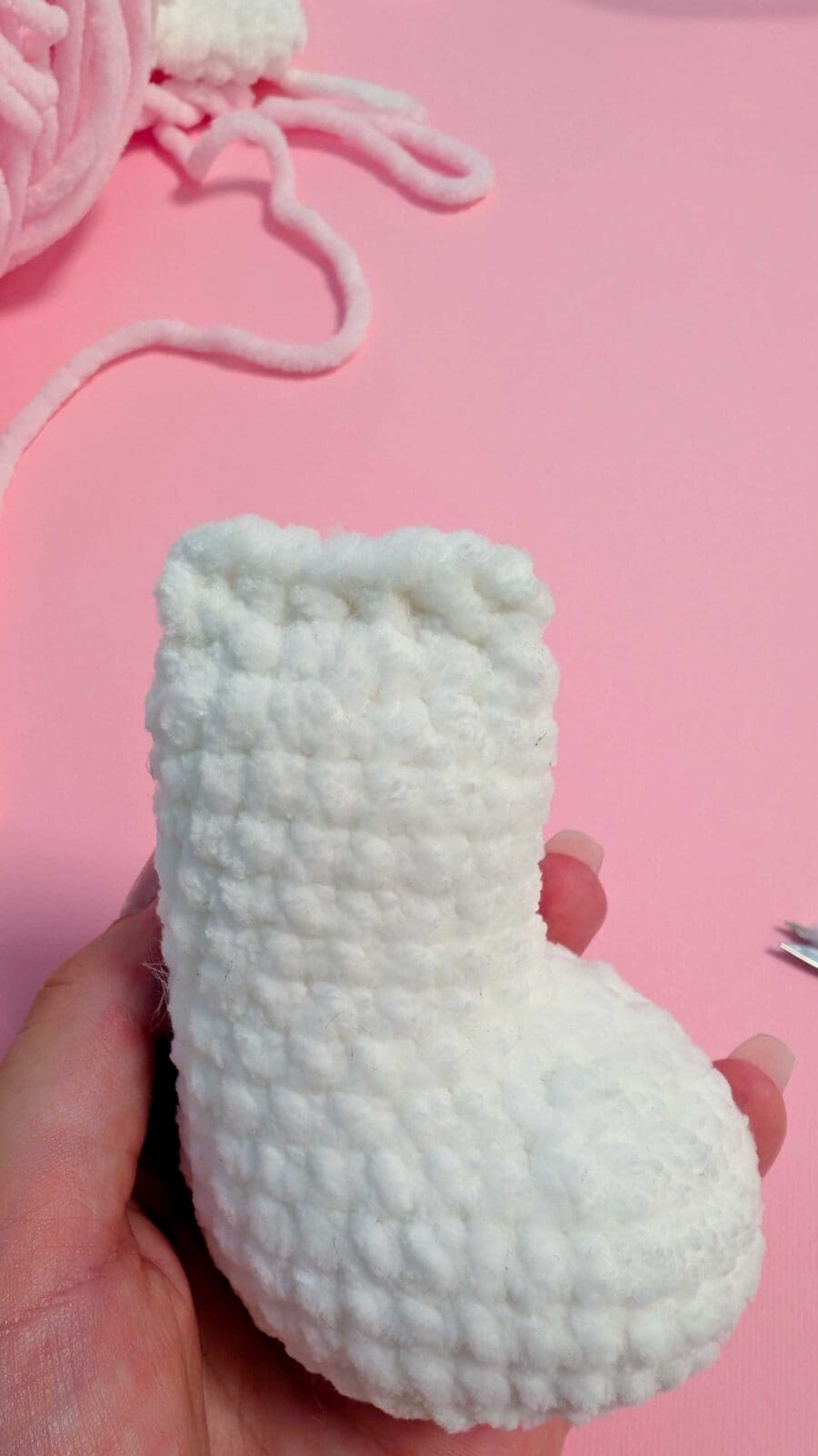 A hand holds a small, white, textured crocheted baby bootie against a pink background with pink yarn and metal crochet tools nearby, perfect for anyone interested in learning how to crochet adorable baby items.