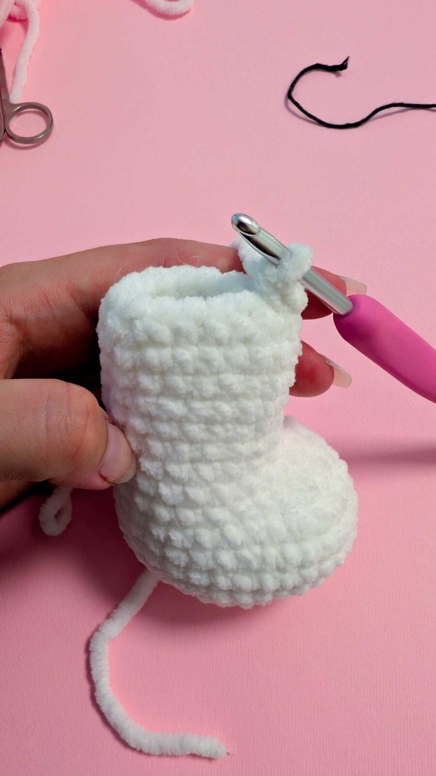 A hand demonstrates how to crochet a small, white baby bootie, using a pink crochet hook to skillfully work with the yarn against a light pink background.