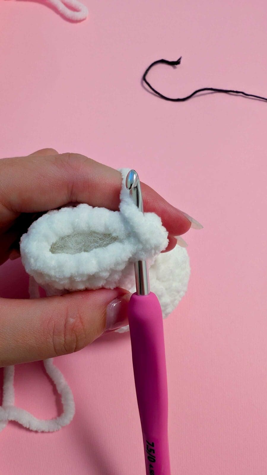 A hand demonstrates how to crochet, holding a piece of white fluffy yarn fabric while a pink-handled crochet hook is inserted into a stitch. The background is pink, with a strand of black yarn visible above.