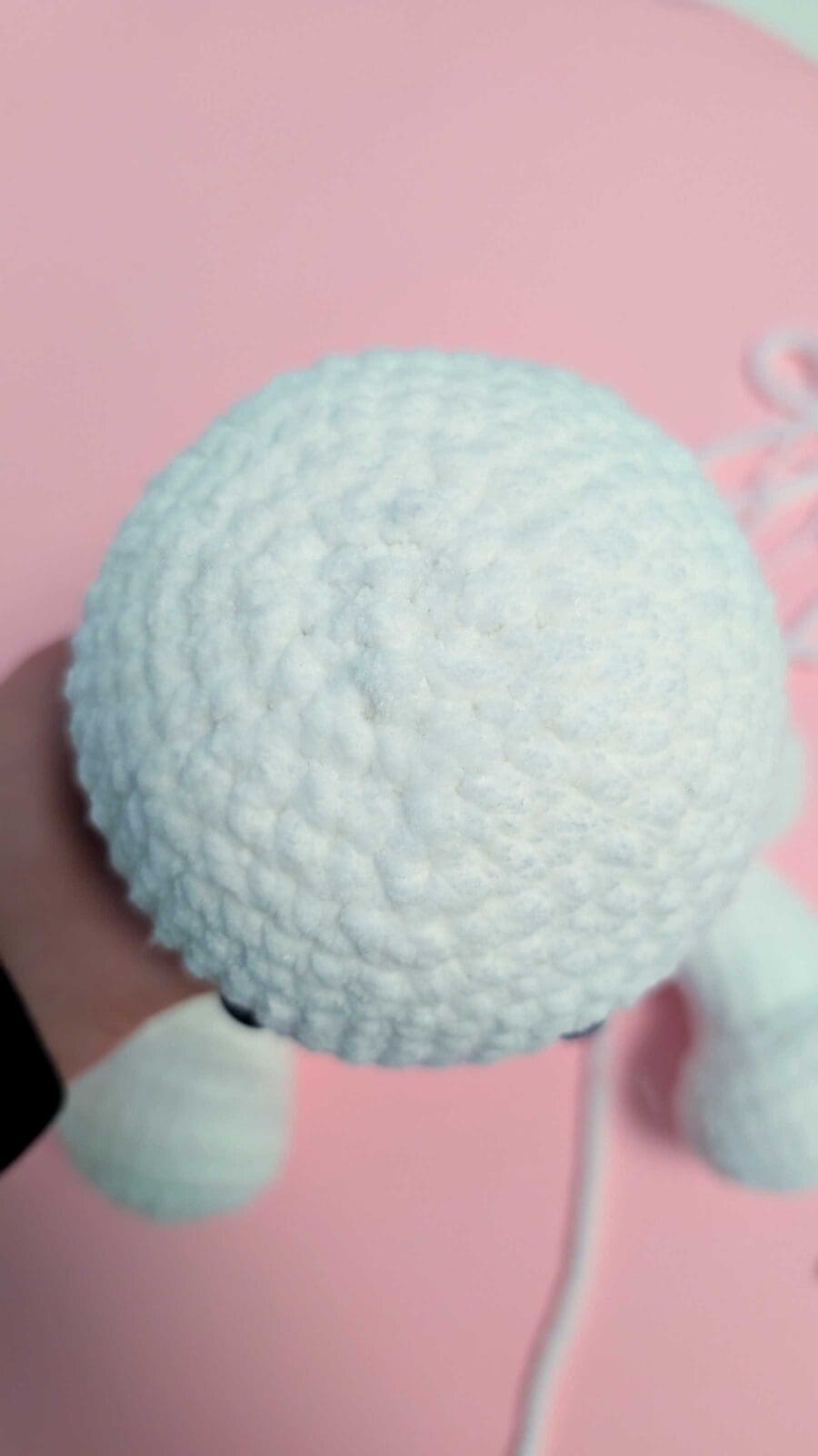 A hand holds a round, white, crocheted plush figure with two small legs visible, set against a pink background with white yarn nearby—a perfect inspiration for anyone learning how to crochet.