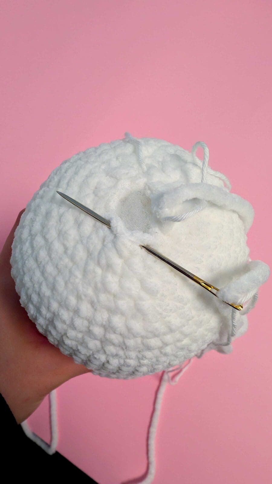 A hand demonstrates how to crochet by holding a white yarn ball with a needle inserted into the stitches, against a pink background. The yarn tail and some loose stuffing are visible at the opening.