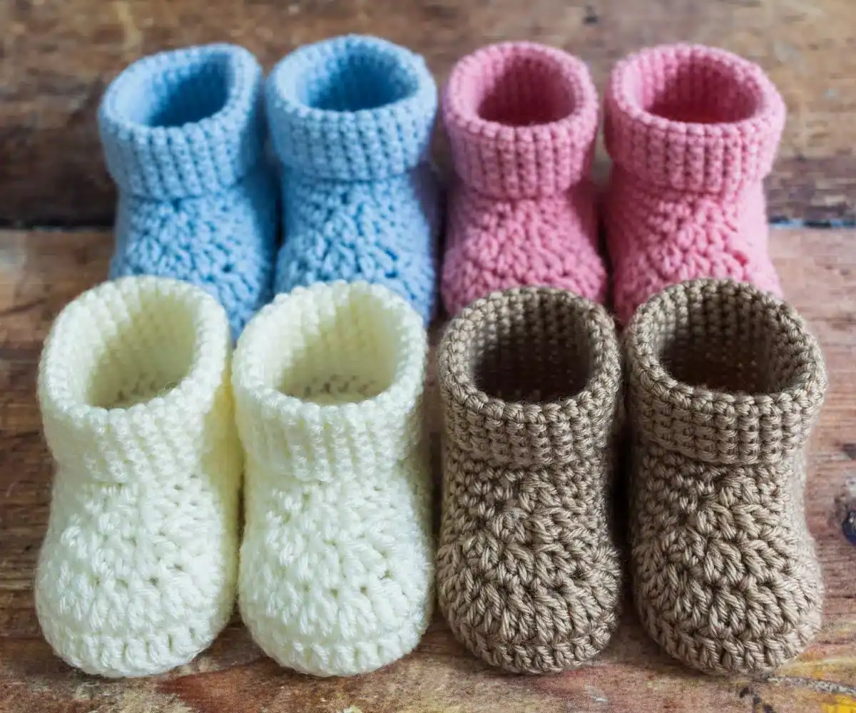 Four pairs of crocheted baby booties in blue, pink, yellow, and brown are neatly arranged on a wooden surface.