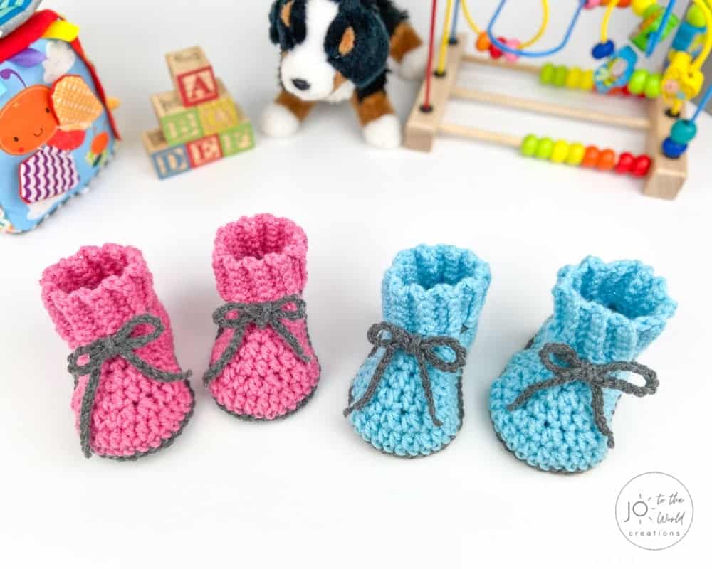 Two pairs of crocheted baby booties, one pink and one blue with gray ties, are displayed on a white surface. In the background, there are colorful toys, building blocks, and a plush dog.