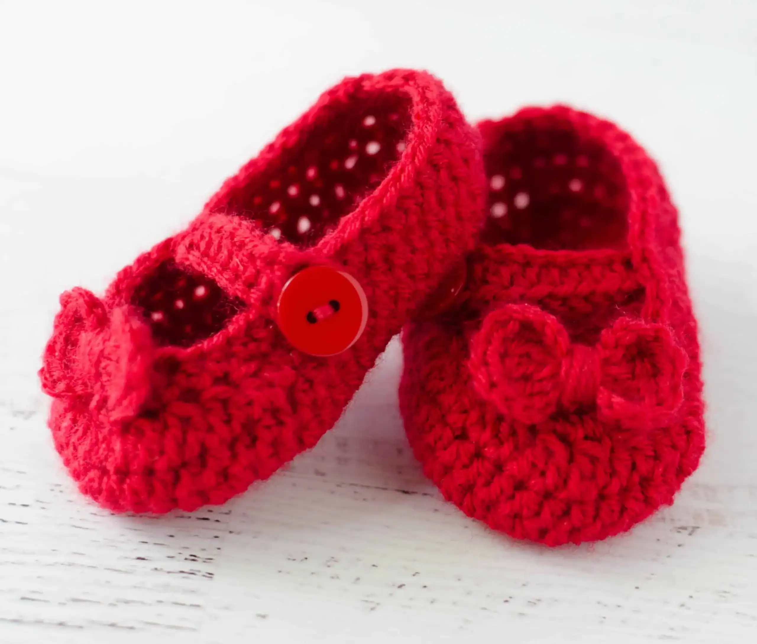 Two crocheted red baby booties with bows and red buttons, placed on a white wooden surface.