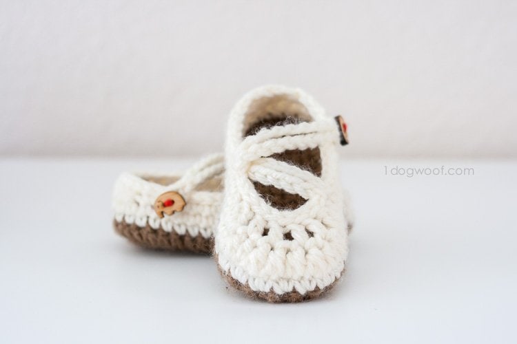 A pair of handmade baby shoes, crocheted with cream-colored yarn and brown soles, featuring small red ladybug buttons on the straps, displayed on a white surface.