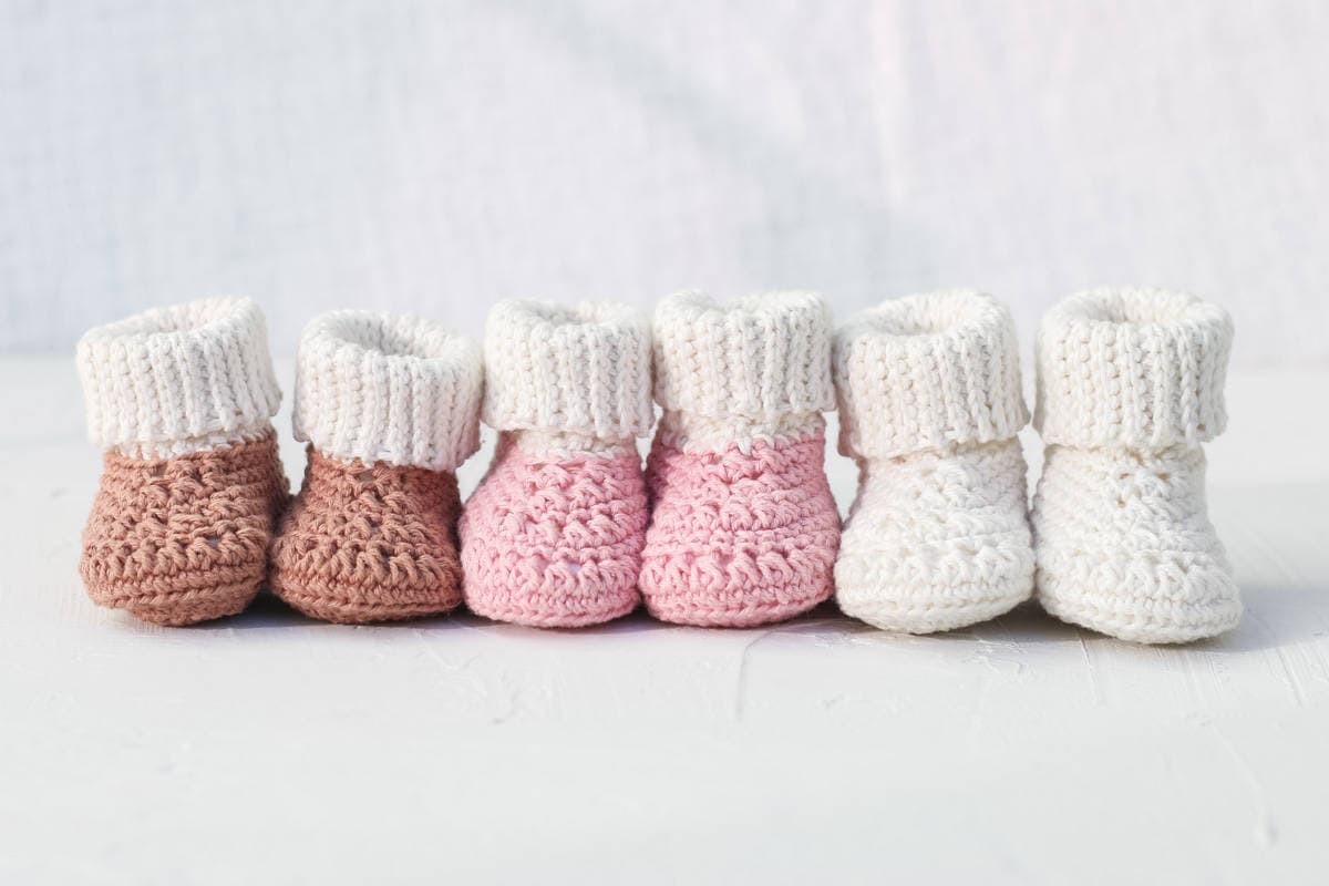 Three pairs of crocheted baby booties in brown, pink, and white are neatly lined up on a light surface against a soft, white background.