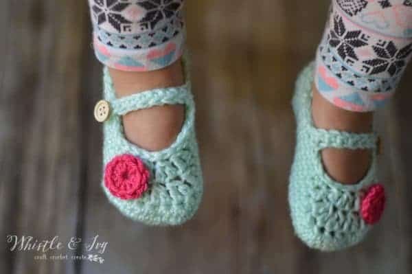 A child wearing pastel patterned leggings and light green crocheted baby booties with pink flower decorations and button straps, standing on a wooden surface.