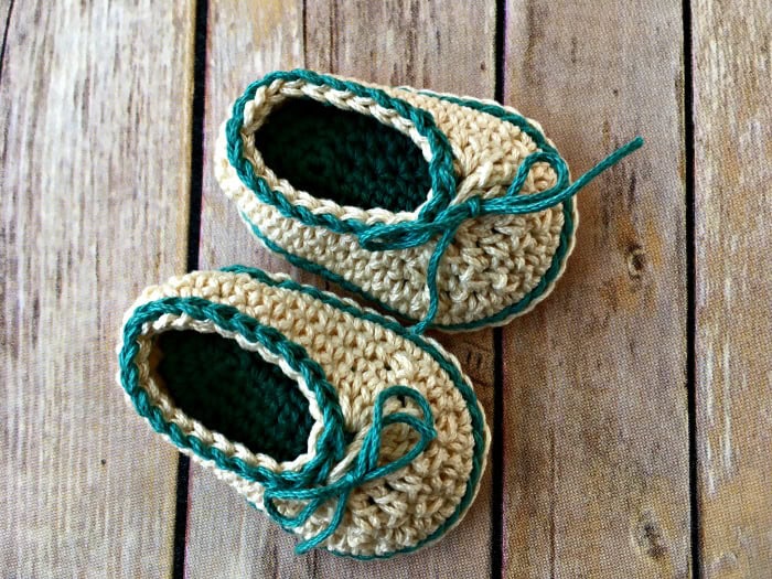 A pair of hand-crocheted baby shoes in beige with teal trim and laces, placed on a wooden surface.