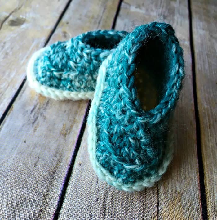 A pair of handmade crochet baby shoes in teal and cream colors are placed on a rustic wooden surface.