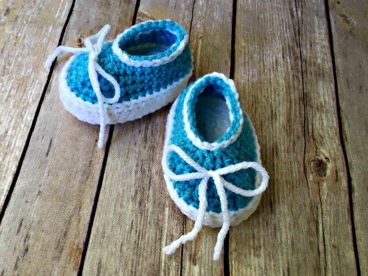 A pair of handmade blue and white crocheted baby shoes with white laces, placed on a rustic wooden surface.