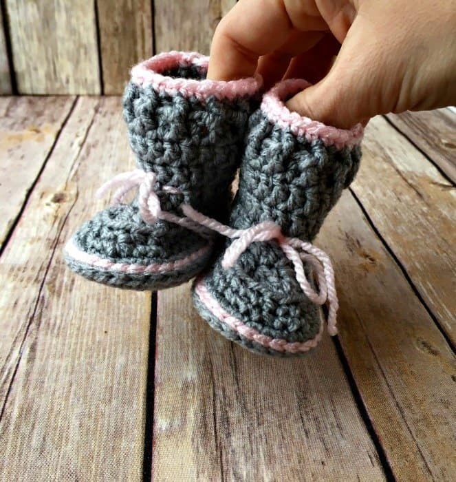 A hand holds up a pair of crocheted baby booties in gray yarn with pink trim and laces, displayed on a rustic wooden surface.