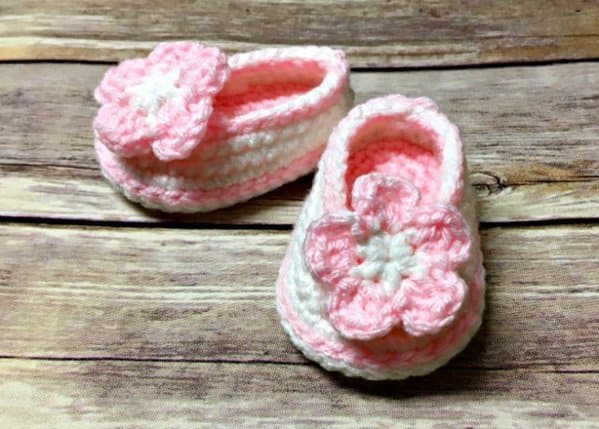 Two crocheted baby booties in pink and white yarn with a large pink and white flower on the top of each, placed on a wooden surface.