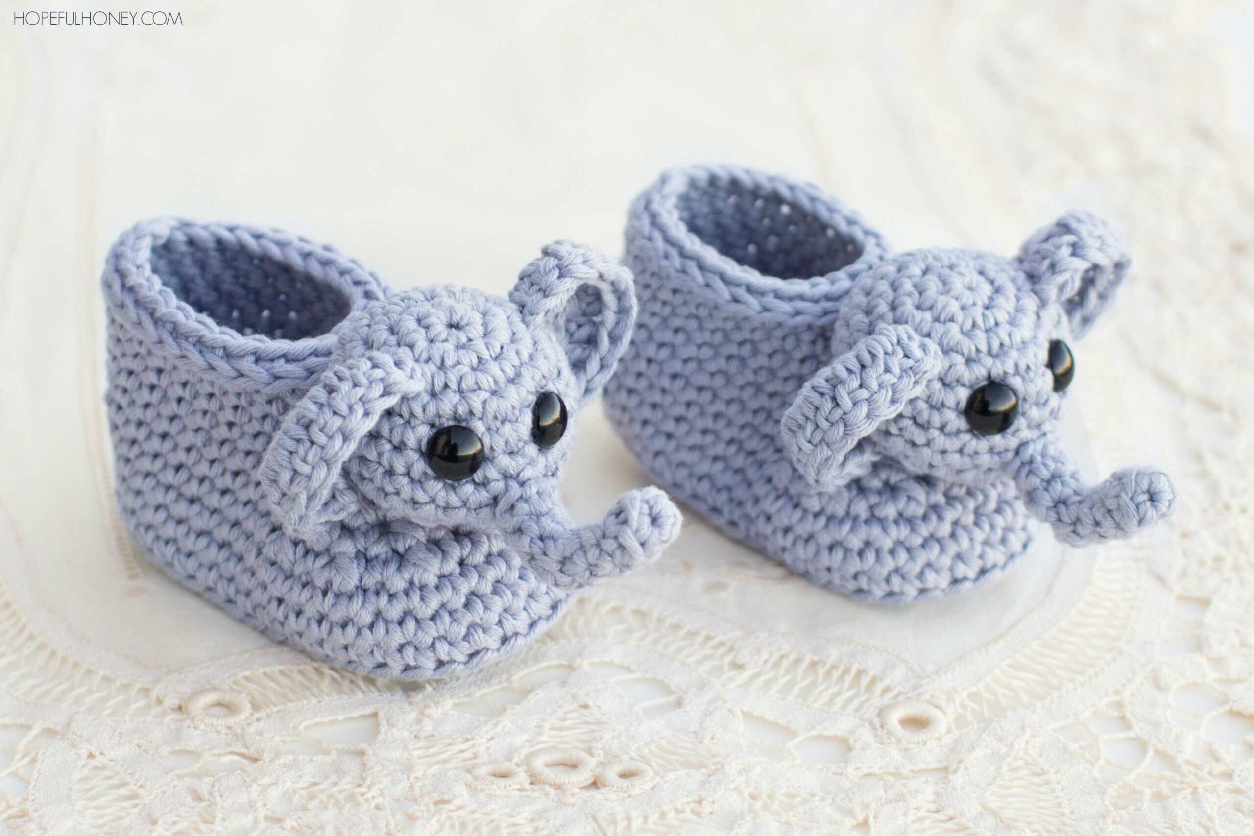 Hand-crocheted baby booties in light purple yarn, designed to look like elephants with protruding trunks, large ears, and black button eyes, displayed on a white lace surface.