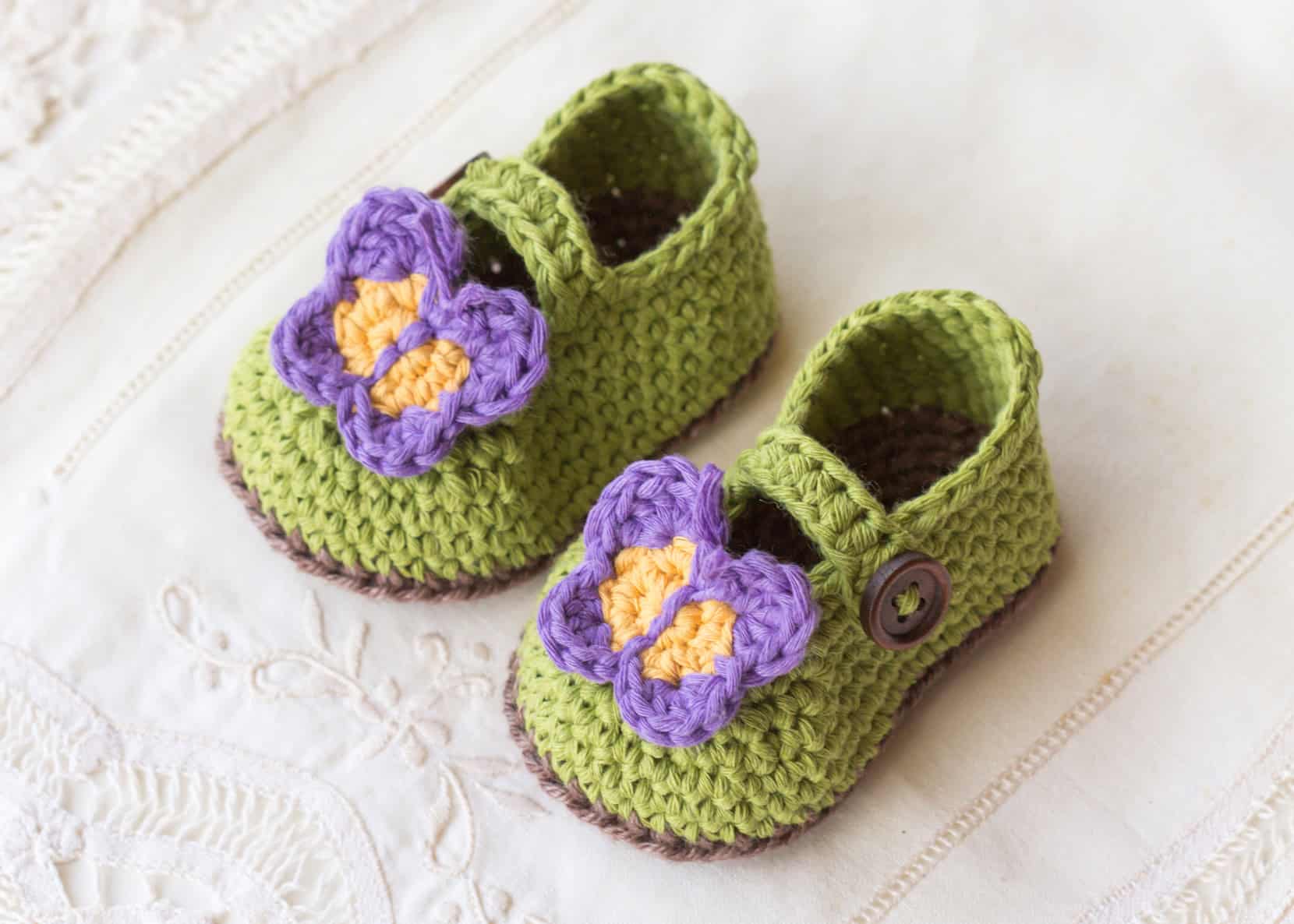 Green baby booties with brown soles, decorated with crocheted purple and yellow butterflies on the toes, placed on a white embroidered fabric.