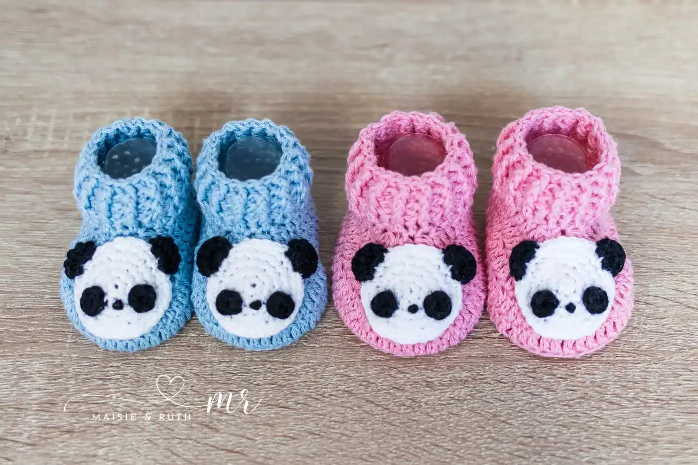 Two pairs of crocheted baby booties, one blue and one pink, each featuring a cute panda face on the front, are placed on a wooden surface.