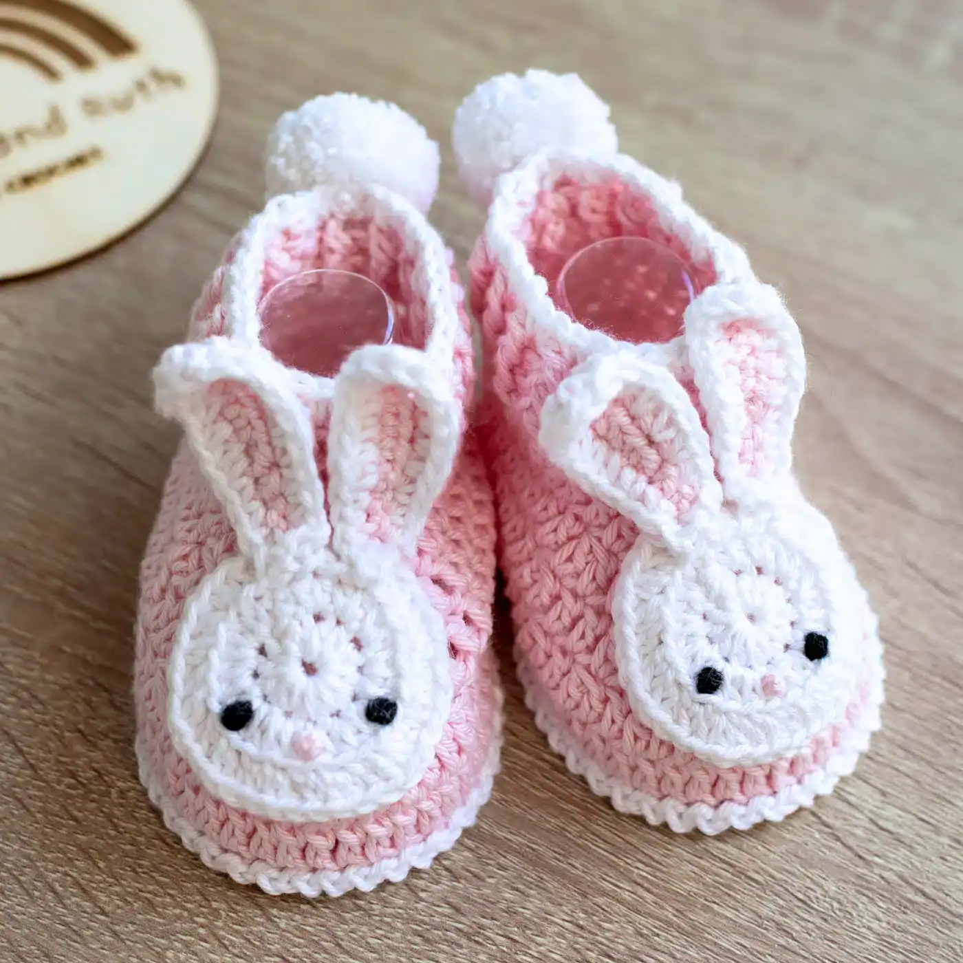 Pink crocheted baby booties with white trim, decorated with white bunny faces and ears on the front. Each bootie has a small white pom-pom on the heel, and they are placed on a light wooden surface.