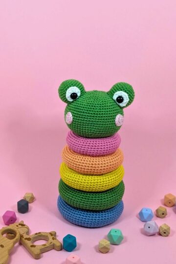 A crochet stacking toy with a green frog head on top and colorful rings below, surrounded by wooden beads and toys, set against a pink background.