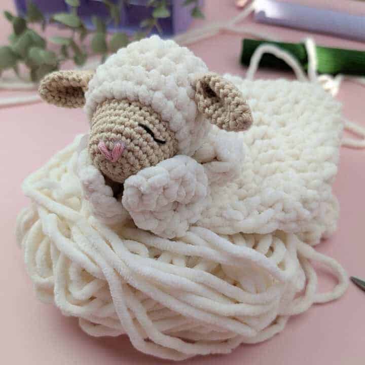 A crochet lamb lovey with closed eyes rests on a coil of soft, white yarn. Greenery and craft supplies are blurred in the pink background.