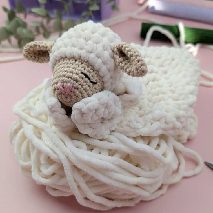 A crochet lamb lovey with closed eyes rests on a coil of soft, white yarn. Greenery and craft supplies are blurred in the pink background.