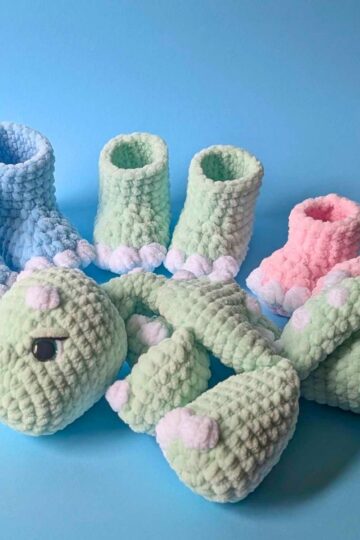 Four pairs of crochet baby booties in blue, green, and pink sit behind a plush green stuffed dinosaur with white and pink accents, all on a light blue background.