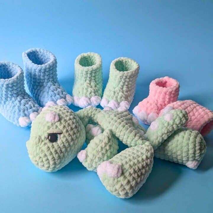 Four pairs of crochet baby booties in blue, green, and pink sit behind a plush green stuffed dinosaur with white and pink accents, all on a light blue background.