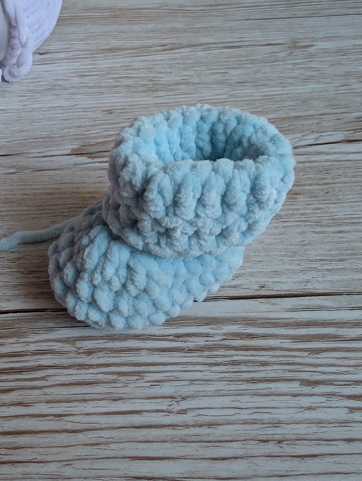 A soft, hand-knitted baby bootie made of light blue yarn sits on a rustic wooden surface.
