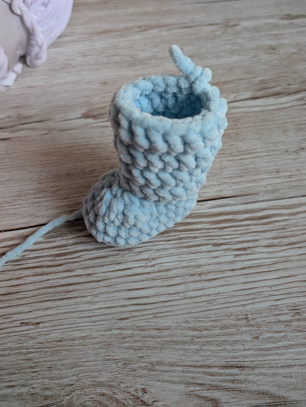 A small, light blue, knitted baby bootie sits on a wooden surface with a loose yarn tail extending from it.