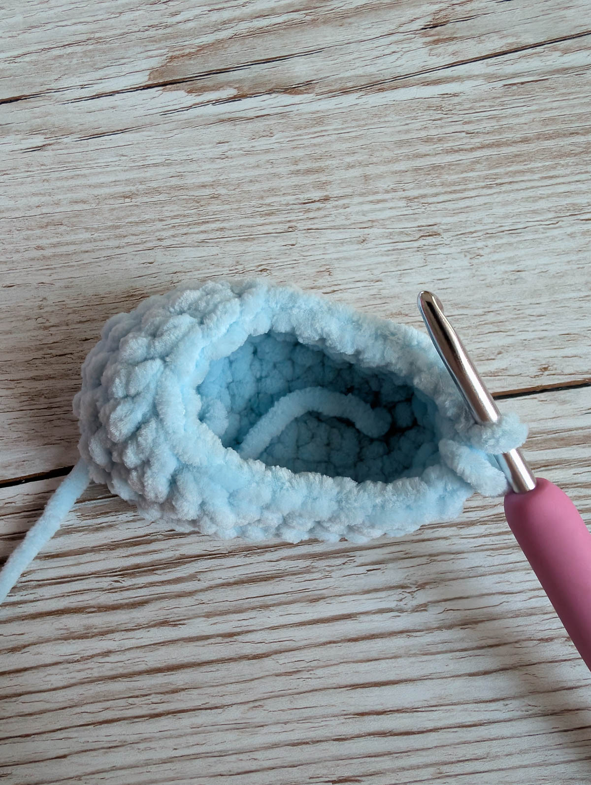 A close-up of a soft, light blue crocheted item in progress, with a pink-handled crochet hook inserted, resting on a light wood surface.
