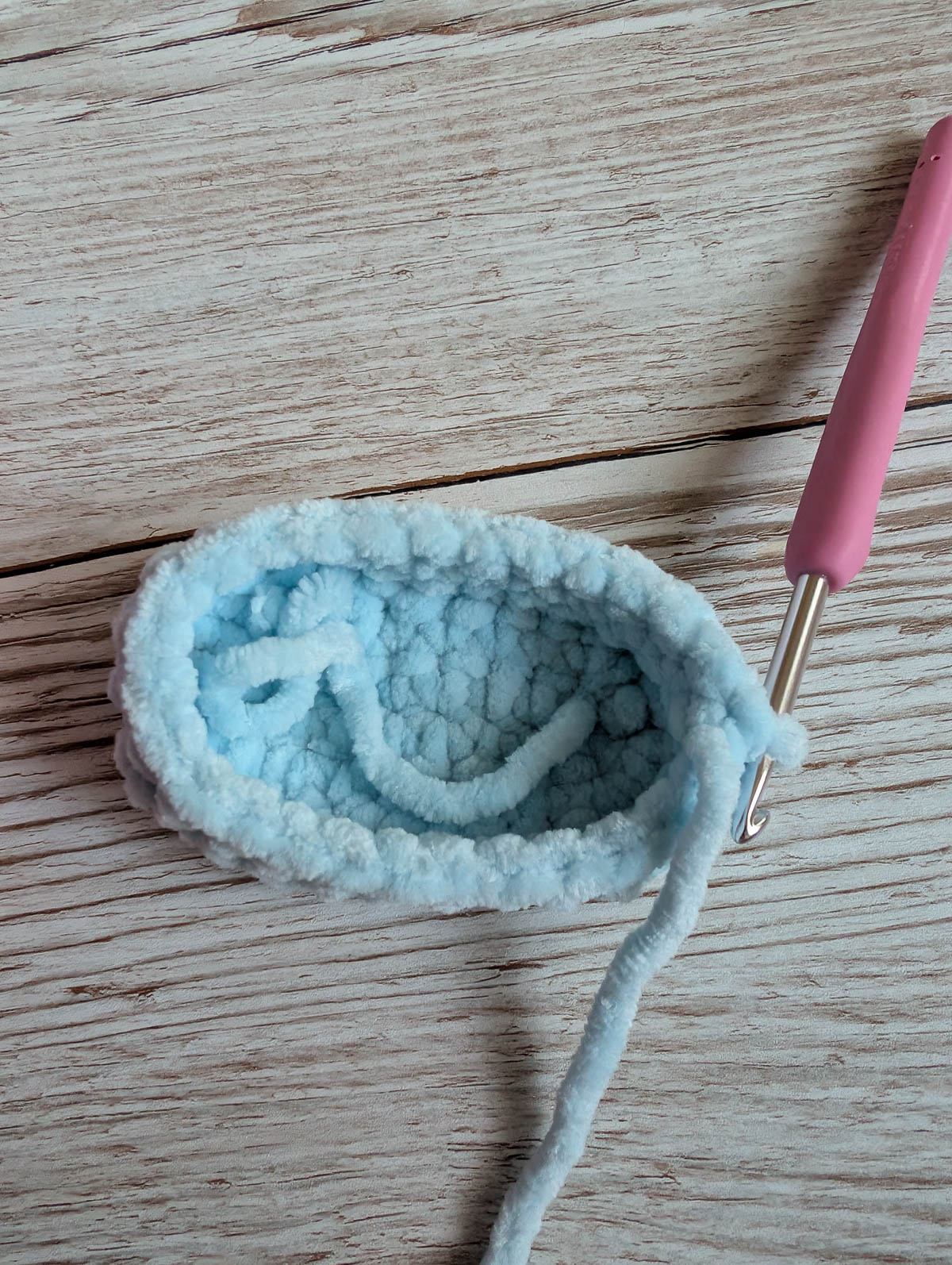 A small, light blue crocheted item in progress, shaped like an oval slipper, rests on a wooden surface. A pink-handled crochet hook is attached to the soft yarn.