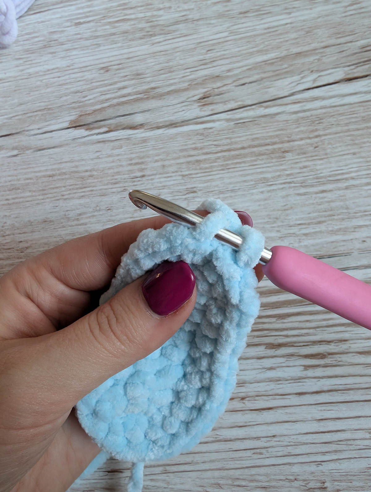 A hand with purple nail polish holds light blue crocheted fabric while using a crochet hook with a pink handle to work through the stitches. The background is a wooden surface.