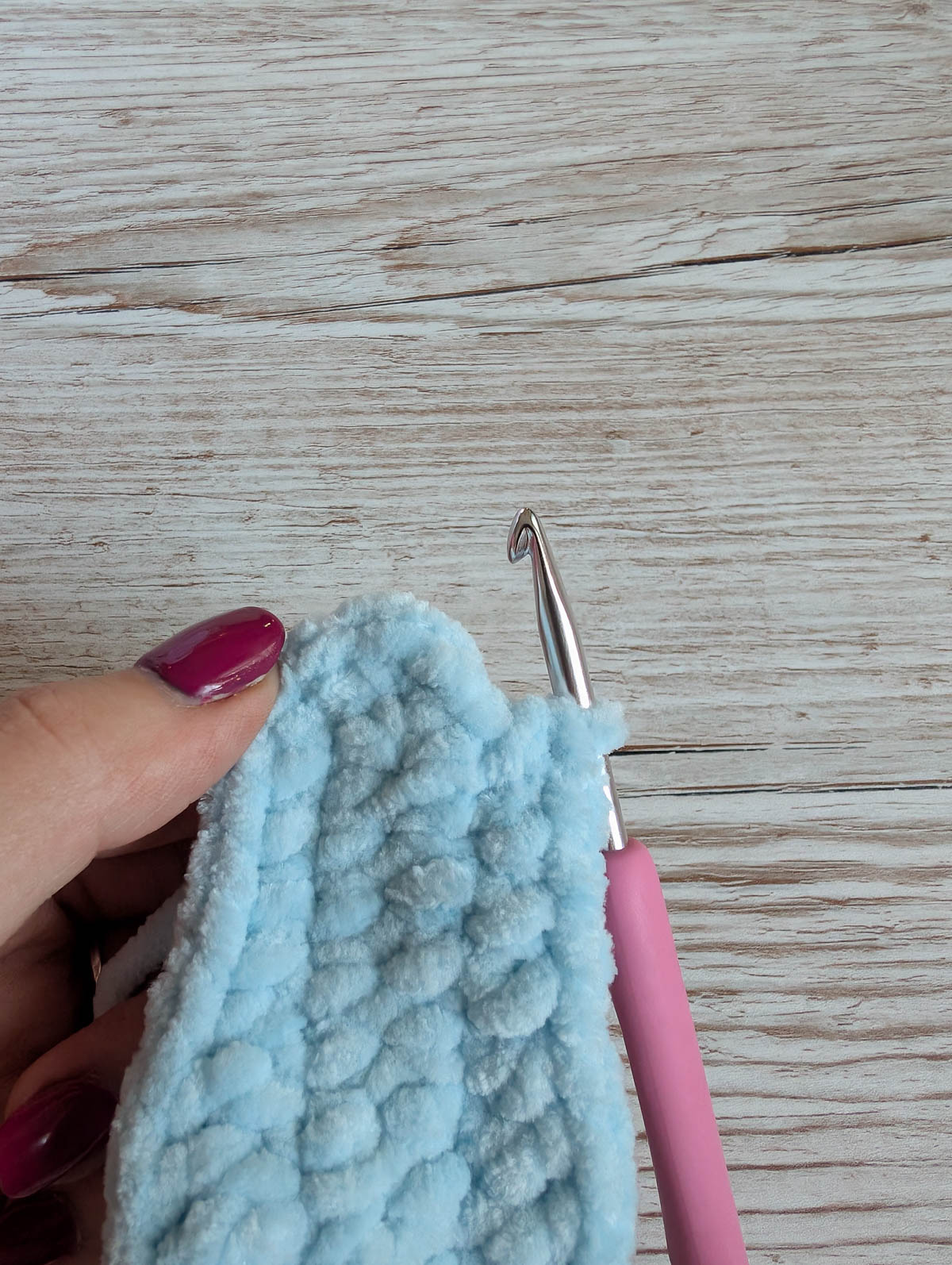 A hand with purple nail polish holds a soft, light blue crocheted piece with a pink-handled crochet hook inserted into the yarn, on a light wooden surface.