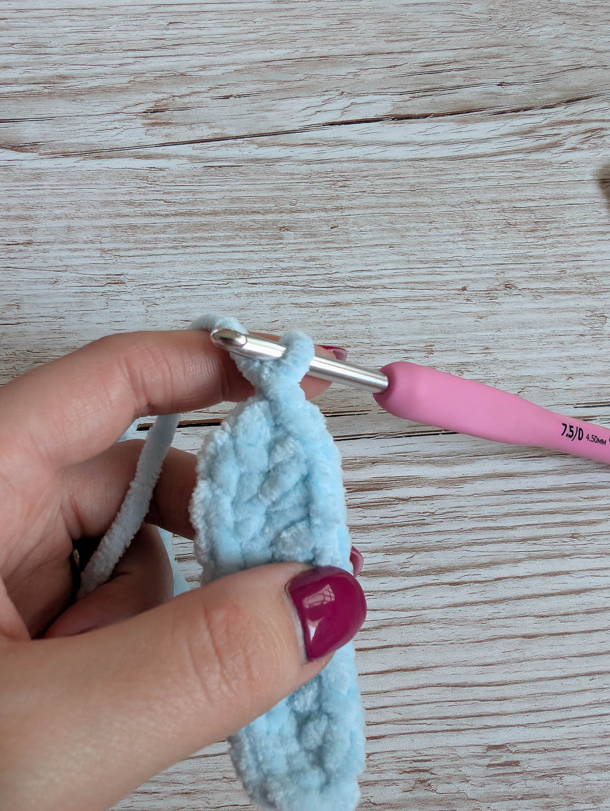 A hand with purple nail polish holds light blue yarn and a pink crochet hook, working on a small, oval crochet piece on a wooden surface.