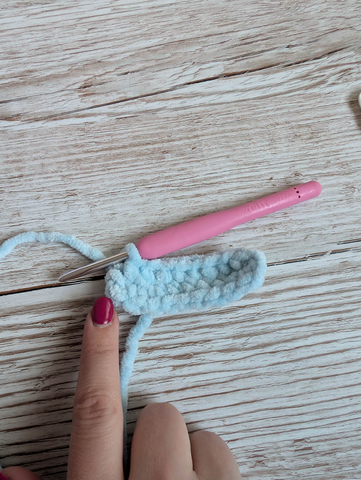 A hand with pink-painted nails points at a small, light blue crocheted piece with a pink crochet hook resting on it, all placed on a light wooden surface.