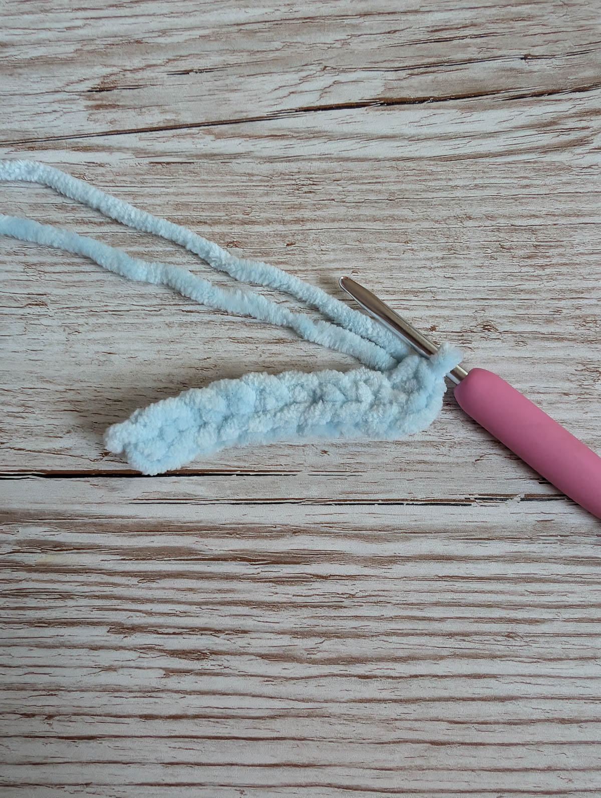 A pink-handled crochet hook holds light blue chenille yarn, partially crocheted, on a light wooden surface.