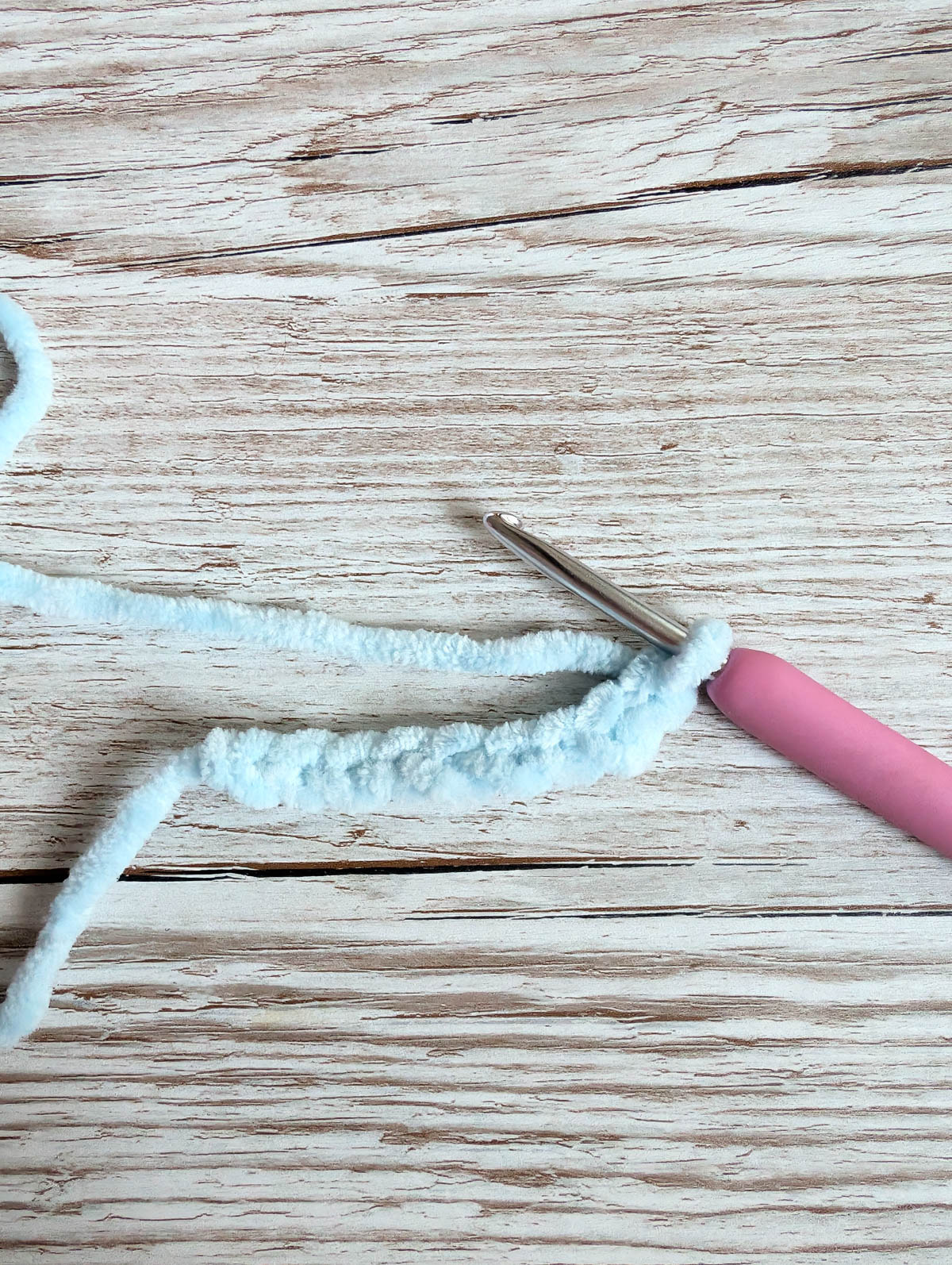 A pink-handled crochet hook with white yarn forms a short crocheted chain, resting on a light wooden surface.