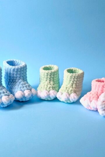 Three pairs of soft, knitted baby booties in pastel colors—blue, green, and pink—are arranged in a row on a light blue background. Each pair features little white toe details resembling bear paws.