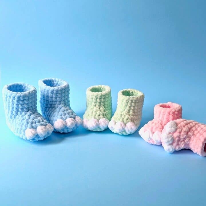 Three pairs of soft, knitted baby booties in pastel colors—blue, green, and pink—are arranged in a row on a light blue background. Each pair features little white toe details resembling bear paws.