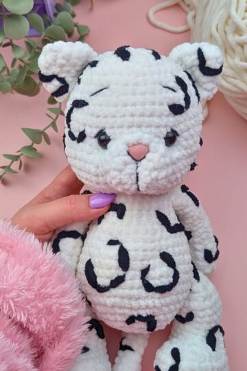 A hand holding a crocheted plush toy resembling a white leopard with black spots. The background includes green leaves, white yarn, a purple box, and a pink fuzzy sleeve, all on a pink surface.