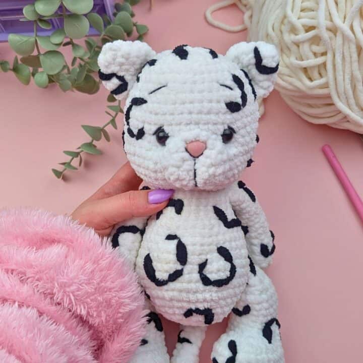 A hand holding a crocheted plush toy resembling a white leopard with black spots. The background includes green leaves, white yarn, a purple box, and a pink fuzzy sleeve, all on a pink surface.
