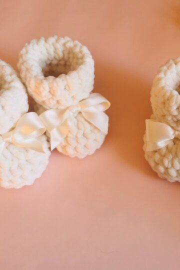 Two pairs of soft, white crochet baby booties with satin bows are displayed on a pale pink surface.