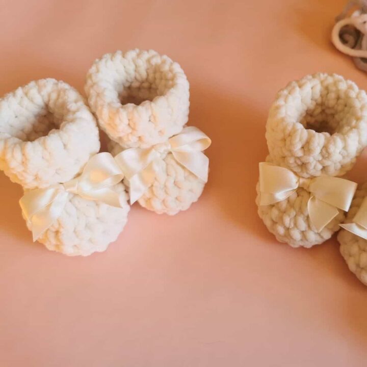 Two pairs of soft, white crochet baby booties with satin bows are displayed on a pale pink surface.