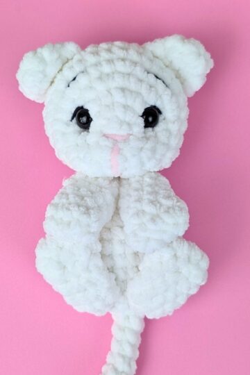 A white, crocheted stuffed animal resembling a cat with black eyes and a pink nose lies on a pink background.