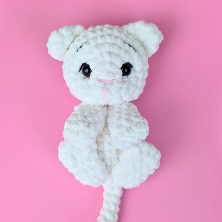 A white, crocheted stuffed animal resembling a cat with black eyes and a pink nose lies on a pink background.