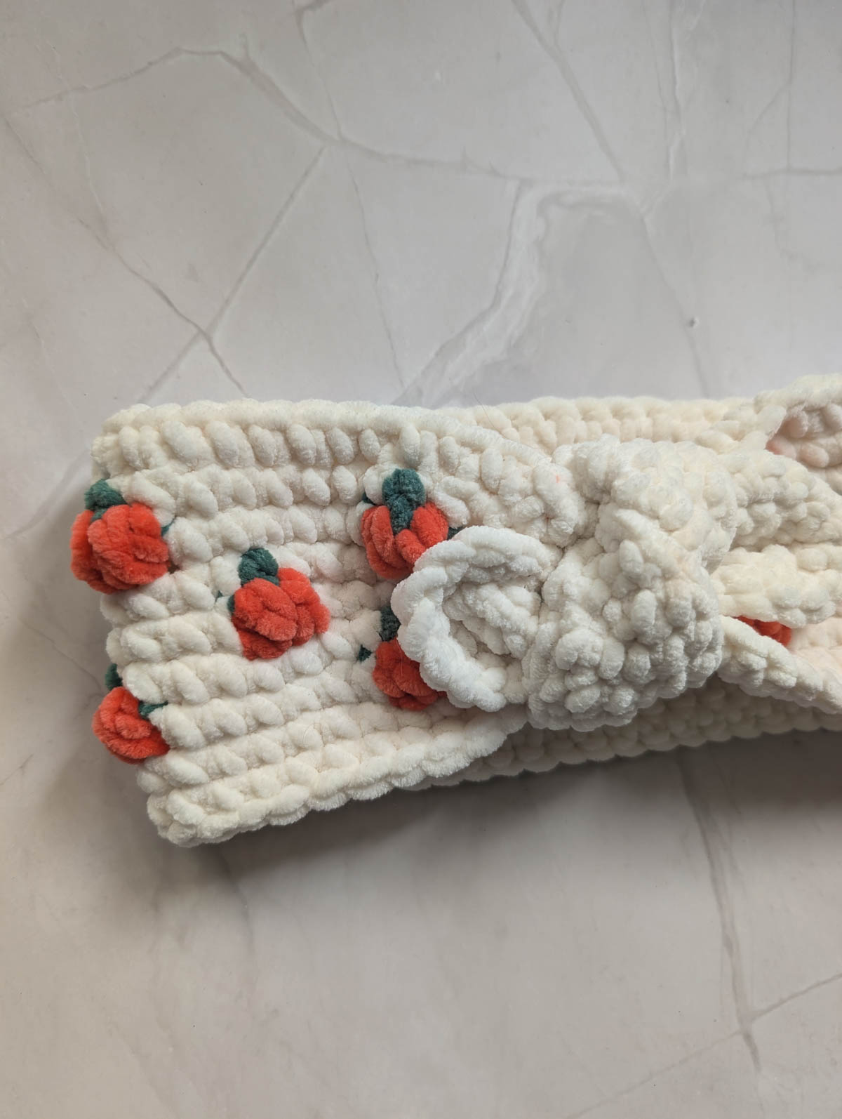 A soft, white, crocheted pumpkin headband with small orange and green pumpkin decorations and a central knot, placed on a light-colored, textured surface—perfect for those seeking a charming crochet pattern.