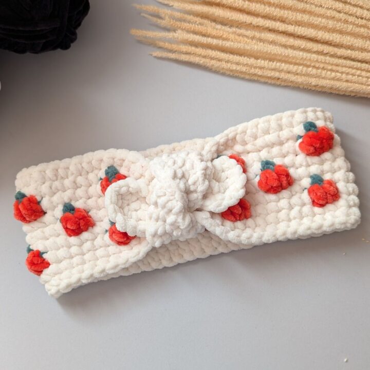 A white crocheted headband with red and green embroidered berry designs and a central knot, displayed on a light surface—this charming accessory is inspired by our popular pumpkin headband and includes a free crochet pattern for creative makers.
